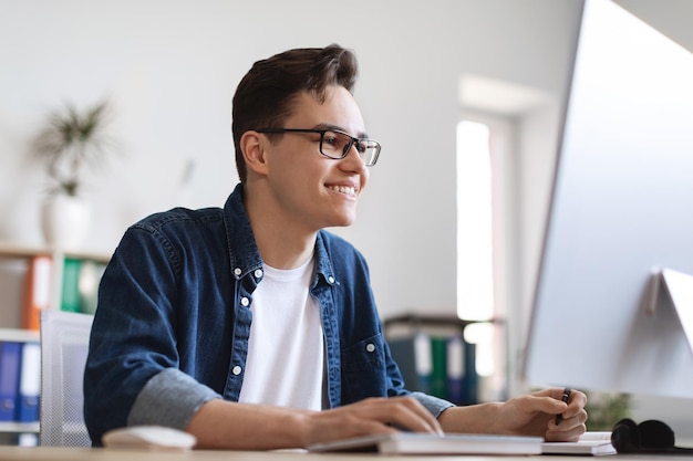 Photo young male office employee taking notes while working on computer at desk