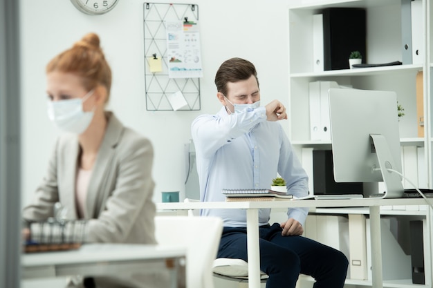 Фото Молодой мужской офис-менеджер в защитной маске, закрывая лицо рукой во время чихания на рабочем месте с коллегой-женщиной, сидящей впереди