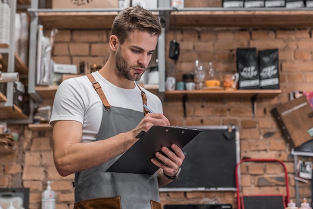 Young male owner writing on clipboard while checking products\
at coffee shop