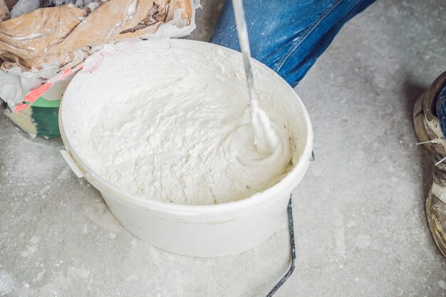 Young male painter kneads putty with water in a bucket using a handheld mixer for building mixes