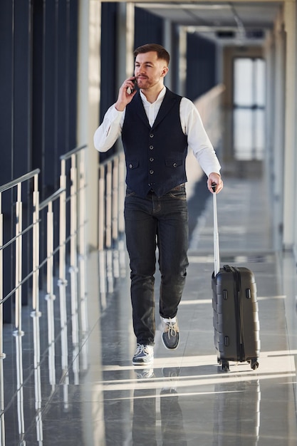 Young male passenger in elegant formal clothes is in the airport hall walking with baggage and talking by phone.