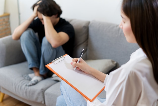 Photo young male patient sitting on sofa with sad face is having consultant by professional psychologist female.