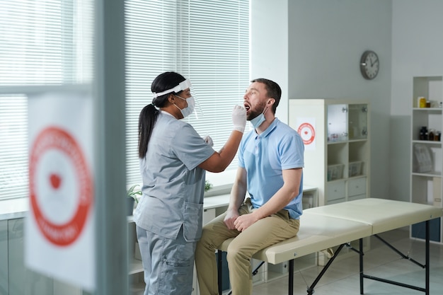 Young male patient with open mouth sitting in front of female clinician in protective workwear and being tested for covid with oral swab in clinics