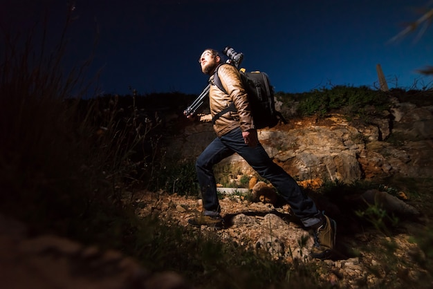 Young male photographer walking on rock beach with camera on tripod in the evening