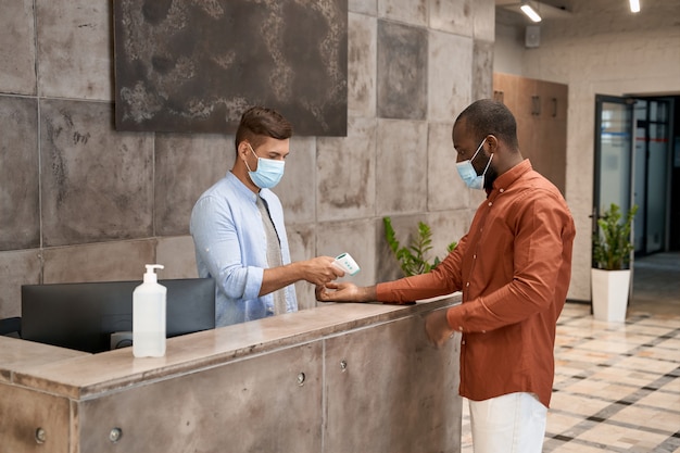 Giovane receptionist maschio che indossa una maschera medica in piedi alla reception nell'ufficio moderno e