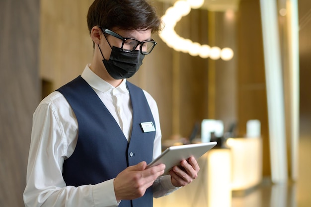 Молодой портье современного отеля в форме и маске с помощью планшета