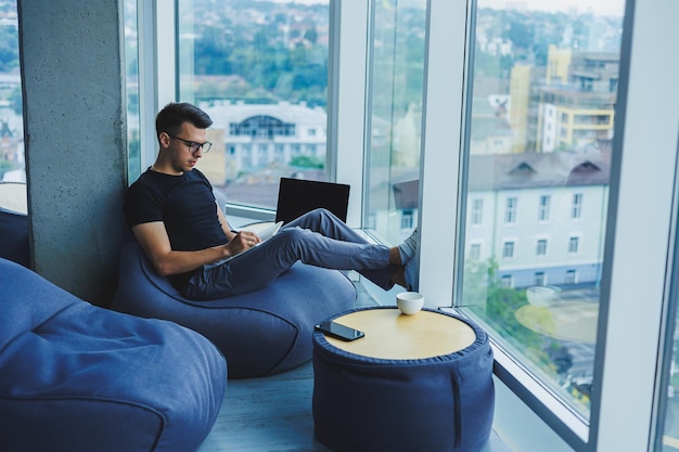 A young male student of caucasian appearance sits in a bag\
chair and takes notes in a notebook the manager sits by the window\
and writes the work schedule