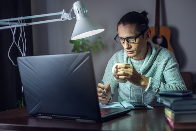 A young male student is studying in an online lesson using a laptop at night by the light of a lamp in the room