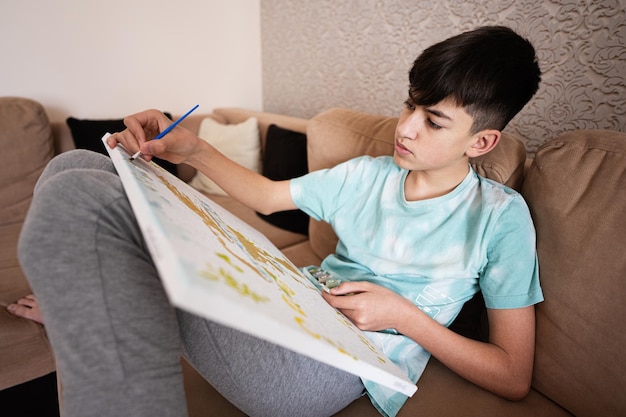 Young male teenager painting picture by numbers on canvas in living room at home Hobby and leisure c