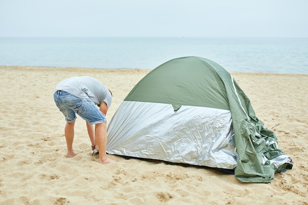 Young male tourist puts a green tent in the beach coast the man sets up a camp on a hike collects a tent alone new normal travel