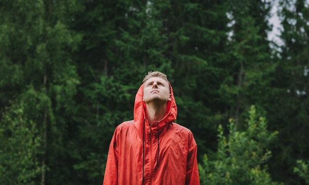 Young male tourist in a red raincoat stands in the rain in the\
mountains against a background trees