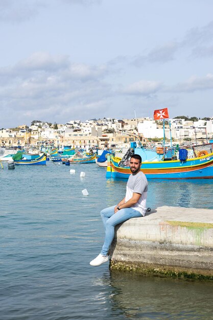 몰타의 Marsaxlokk에서 다채로운 보트로 가득한 바다를 배경으로 앉아 있는 젊은 남성 관광객