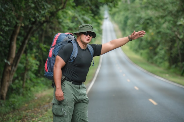 Young male tourists carrying backpack to travel by hitchhiking. Travel by hitchhiking. Tourists wait to hitchhike or wave for help at the roadside in the forest of Khao Yai National Park.