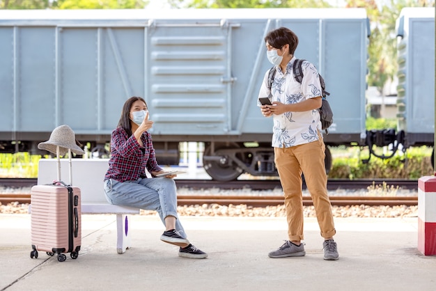 Young male traveler backpack with mask and mobile in hand asking way for help from woman sitting and pointing on stairs at subway, covid distance