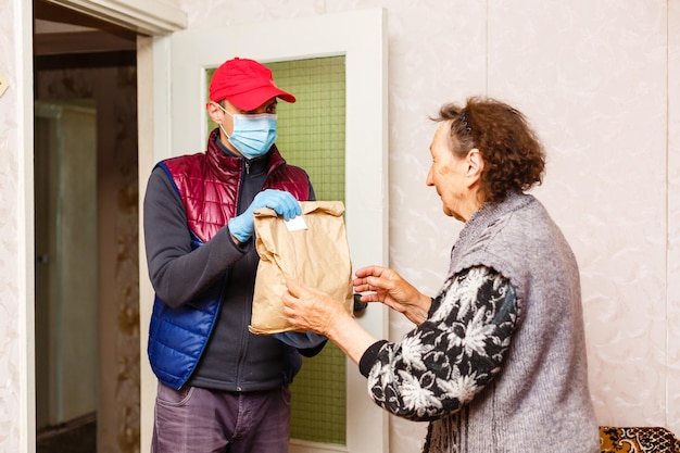Молодой мужчина-волонтер в маске раздает пожилой женщине коробки с едой возле ее дома. Сын-мужчина помогает пожилой матери-одиночке. Поддержка семьи, забота. Карантин, изоляция. Коронавирус (COVID-19. Пожертвование
