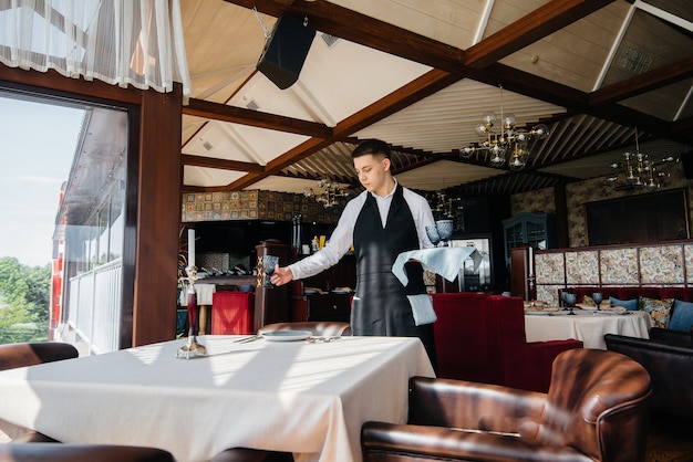 スタイリッシュな制服を着た若い男性ウェイターが、美しいグルメレストランでテーブルを提供しています。最高レベルのレストラン活動。