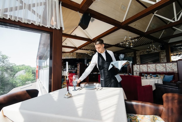 スタイリッシュな制服を着た若い男性ウェイターが、最高レベルの美しいグルメレストランレストランでテーブルを提供しています。