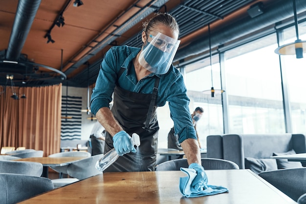 Молодые мужчины-официанты в защитной спецодежде чистят столы в ресторане