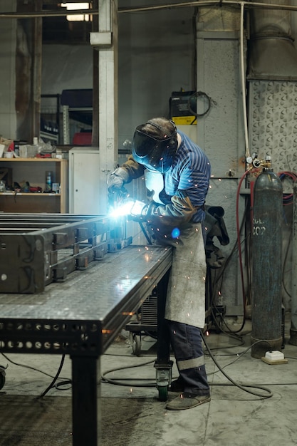 Young male welder in apron and other protective workwear\
working in factory