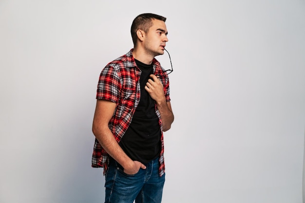 Young male with eyeglasses to lips thinking a deep thought on white background a portrait of handsome man in shirt and jeans with glasses in his hand and mouth concept problem solving