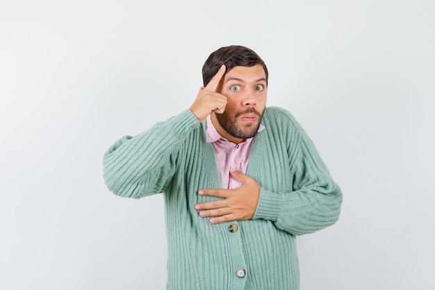 Young male with hand on chest while pointing at head in shirt, cardigan and looking pensive. front view.