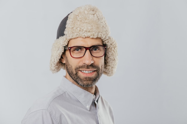 Young male with pleasant appearance, wears warm fut hat, transparent glasses, models over white studio wall