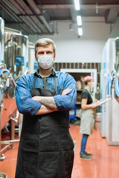 Young male worker of beer production factory in workwear and\
protective mask standing against female colleague rewriting data\
from control panel