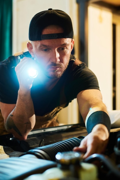 Young male worker or engineer with flashlight checking motor of broken car