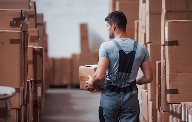 Young male worker in uniform is in the warehouse with box in hands.