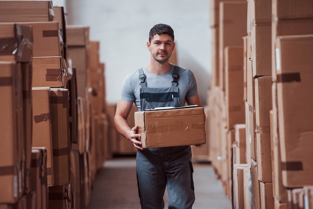 Young male worker in uniform is in the warehouse with box in hands.