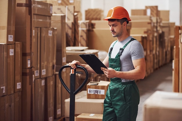 Young male worker in uniform is in the warehouse with notepad and pallet truck.