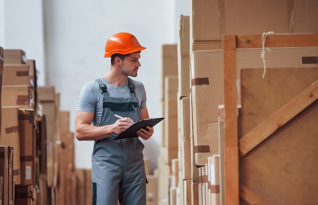 Young male worker in uniform is in the warehouse with notepad and pen.