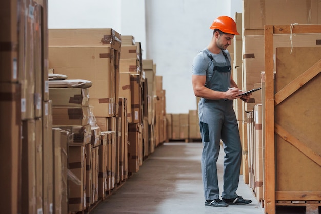 Young male worker in uniform is in the warehouse with notepad and pen.