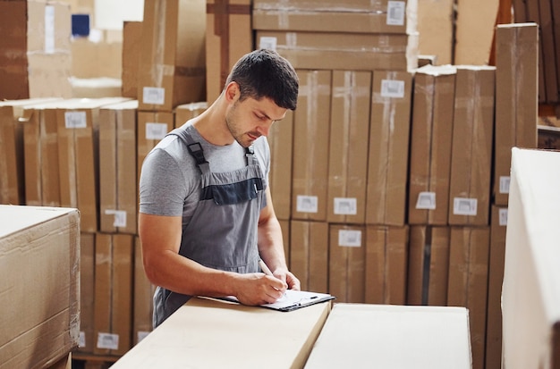 Young male worker in uniform is in the warehouse with notepad and pen.