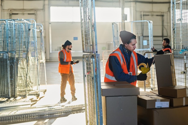 Young male worker of warehouse sealing one of boxes with cellotape