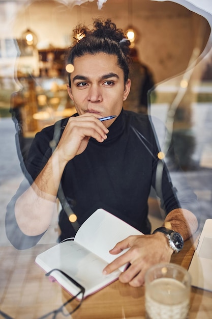 Young male writer with black curly hair sitting indoors in cafe with pen and notepad. View through the glass.