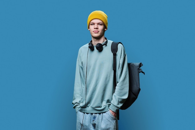 Young male in yellow cap sweatshirt with headphones backpack on blue background