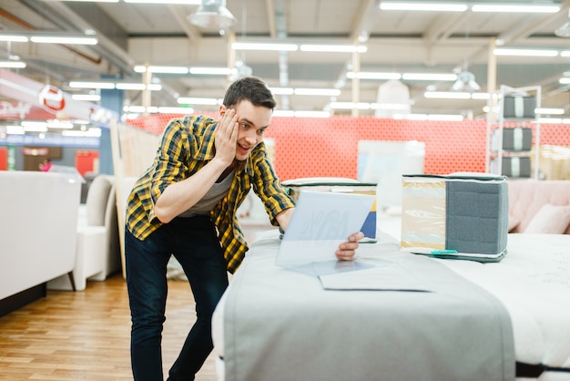 Young man afraids of the price in furniture store showroom. Male person looking samples for bedroom in shop, husband buys goods for modern home interior