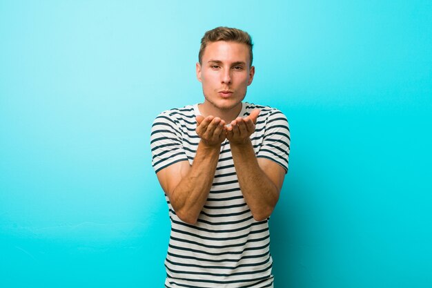 Young man against a blue wall folding lips and holding palms to send air kiss