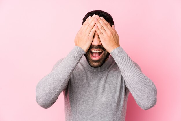 Young man against a pink wall isolated covers eyes with hands, smiles broadly waiting for a surprise