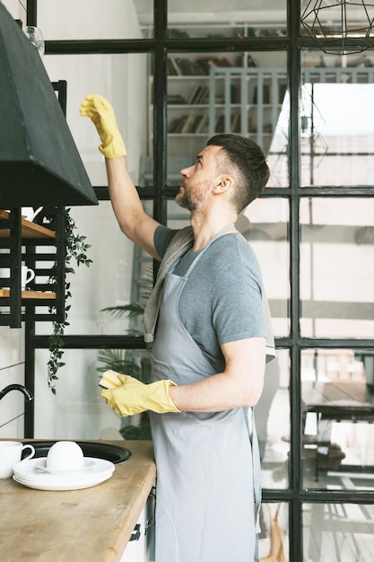 young man in apron and gloves does household chores washes dishes with towel on his shoulder men housework household help in stylish kitchen in modern apartment