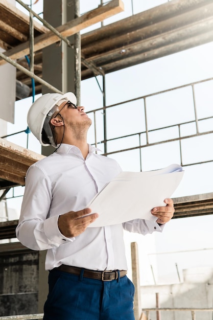 Young man architect or businessman wearing hard hat and holding blueprints on a construction site