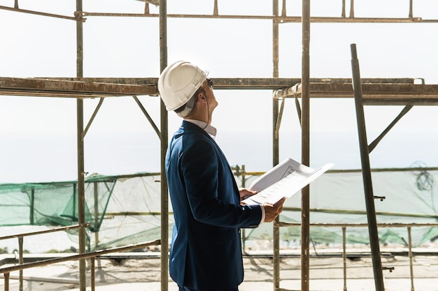 Young man architect wearing formal suit and hard hat during building construction control holding a blueprints on a construction site