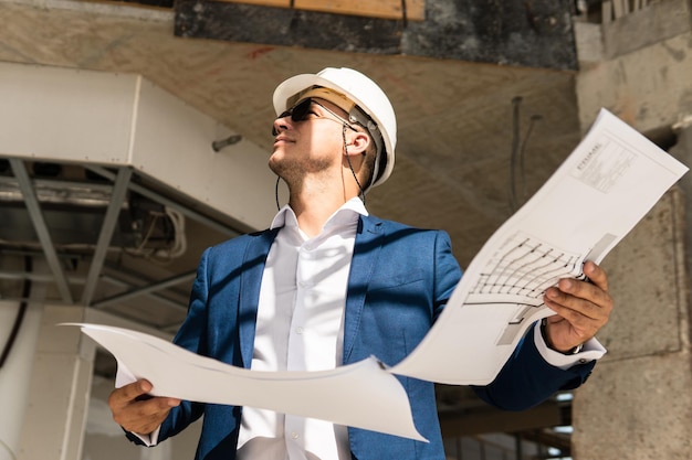 Young man architect wearing formal suit and hard hat during building construction control holding a blueprints on a construction site