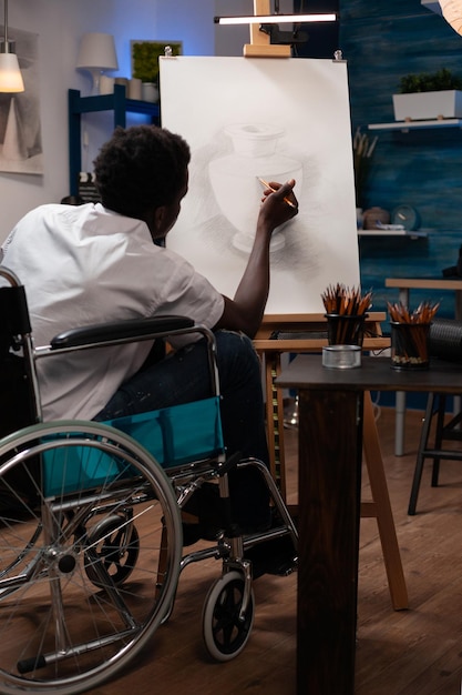 Young man artist in wheelchair making shadow using graphic pencil working at vase ilustration during painting lesson in art studio. Creative student with disability sketching artistic idea