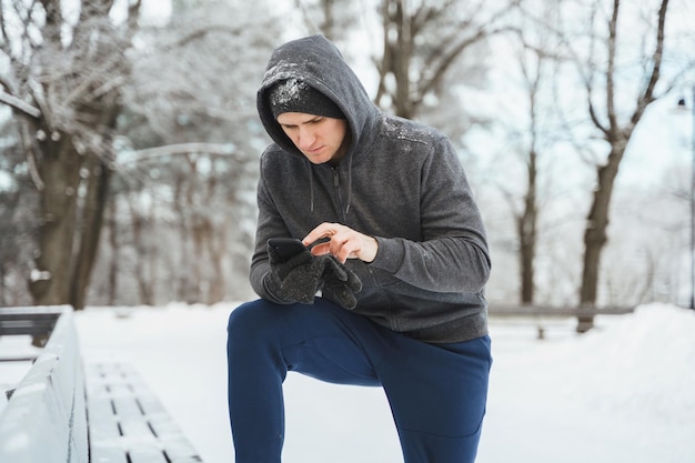 雪の都市公園で冬のトレーニング中にスマートフォンを使用して若い男のアスリート