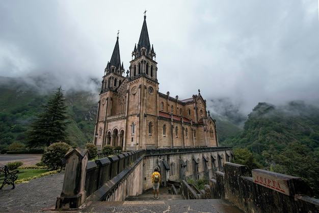 Young man backpacker with a yellow jacket and mask next to the Sanctuary of Covadonga, Santa Maria la Real, on a cloudy day.