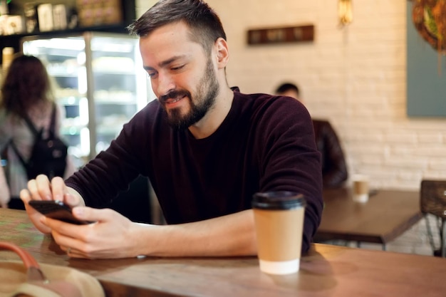 コーヒーブレイクを持ち、モバイルタッチスクリーン電話を使用しているバーの若い男