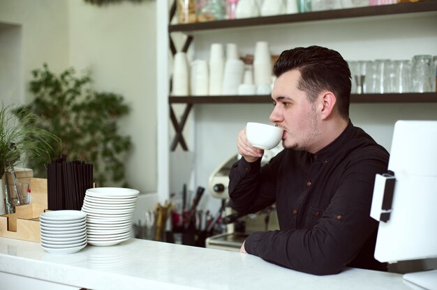 Young man barista in black shirt drinks coffee at the bar