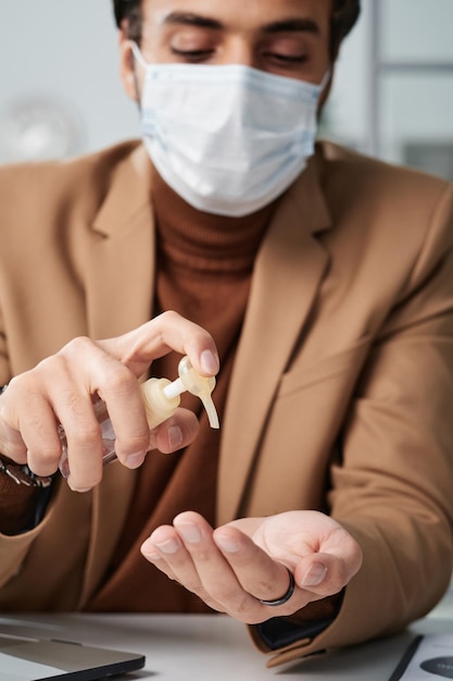 Foto giovane in giacca beige seduto al tavolo e usando un disinfettante per le mani in ufficio